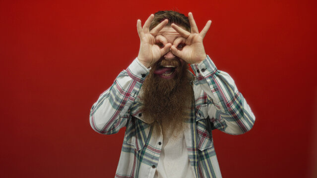 Bearded man wearing plaid shirt making binoculars gesture with tongue out in studio; watching exploring curiosity. - Powered by Adobe