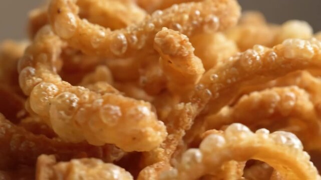 Extreme close-up of crispy chicharrones rotating slowly, revealing puffed golden texture, seasoning flecks, and airy surface details under warm lighting.

