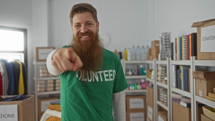 Fototapeta premium Bearded man pointing finger in donation building beside clothes box and shelves; support motivation.