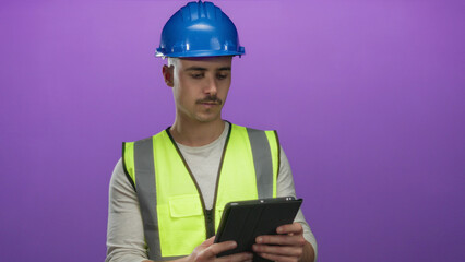 Young man in reflective vest and hardhat examines a tablet against isolated purple background, symbolizing technology and construction with a professional focus.