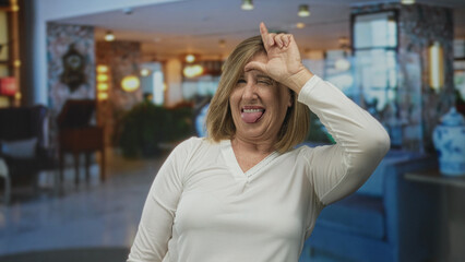 Woman making loser sign with right hand on forehead while smiling in spacious hotel lobby; mockery...