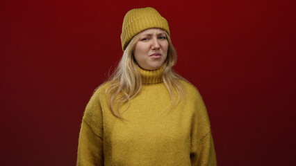 Woman expressing disgust wearing yellow sweater and beanie on red background capturing unique emotion and style.