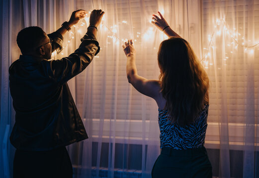A warm scene where two individuals hang decorative string lights on curtains in a dimly lit room, setting a cozy and festive mood. The lighting creates an inviting and joyful ambiance.