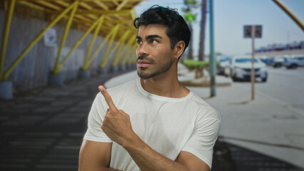 Fototapeta premium Young hispanic man wearing white shirt finger pointing to object on sunny city street near parked cars; curiosity.