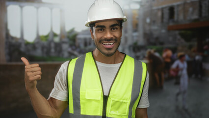 Hispanic man wearing hardhat and reflective vest gives thumbs up gesture at ancient roman building;...