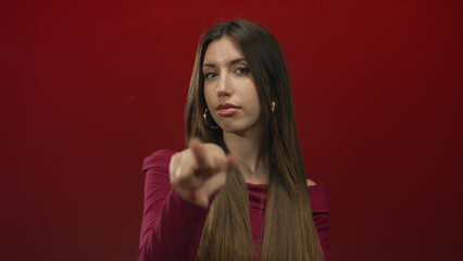 Young hispanic woman with long straight hair points finger forward in red studio setting; alertness.