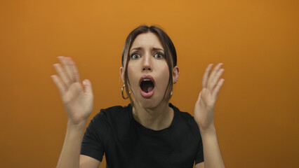 Young hispanic woman with wide eyes showing open mouth and raised palms and hoop earrings in orange...