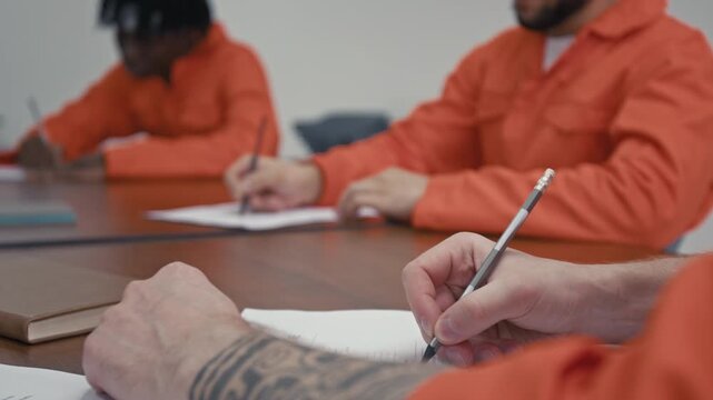 Medium close-up of diverse male criminal convicted offenders in orange jumpsuits learning to write resume during adaptation course in jail, developing social skills before reintegration into society