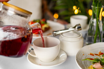Glass teapot pouring red tea into a white cup on a cozy dining table with food and green plants in the background, creating a warm morning meal atmosphere.