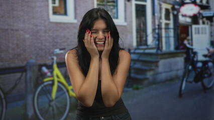 Young hispanic woman cups cheeks with hands while standing near bicycle on street in front of building; joy.
