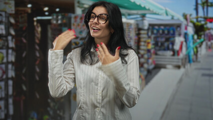 Fototapeta premium Hispanic woman with dark glasses adjusts collar on white striped blouse and smiles in street; self esteem empowerment confidence.