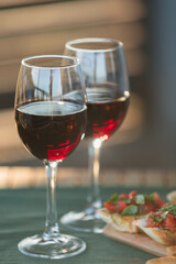 Close-up of red wine glasses and bruschettas with tomatoes and basil on wooden plate on a sunny garden party table.