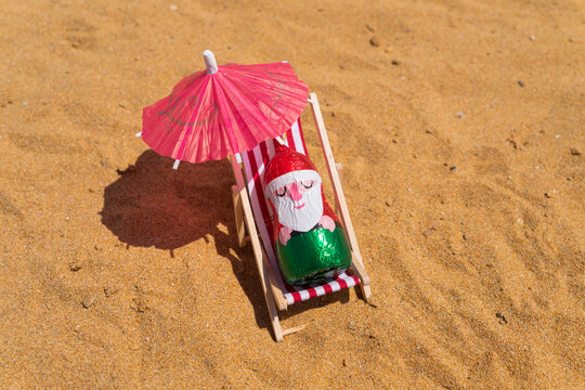 Chocolates Santa sits upright on beach chair, figure shaded by wide paper umbrella. Christmas in July concept