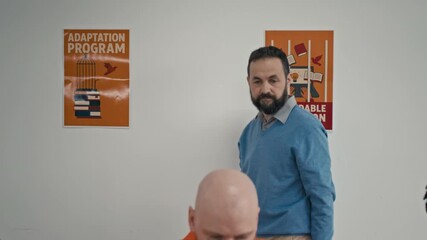 Zoom-out shot of Caucasian male prison teacher hanging up motivational jail poster on wall, then standing behind and observing multiethnic convicts performing tasks on laptops during adaptation course