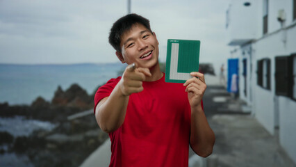 Young man on seaside promenade holding learner sign outdoors with a confident smile and index finger pointing towards the camera against a scenic coastal backdrop.