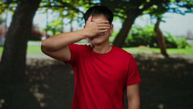 Young man in red shirt covers his eyes with hand while standing in a sunny park surrounded by lush green trees, creating a playful outdoor atmosphere.