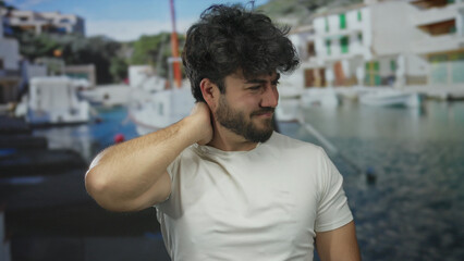 Fototapeta premium Young man with beard experiencing neck discomfort at a picturesque port with boats and ships in the background, possibly depicting stress or pain outdoors.