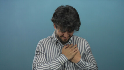 Young hispanic man with beard clutching chest against blue background in a moment of pain, suggesting a heart condition or emotional distress.