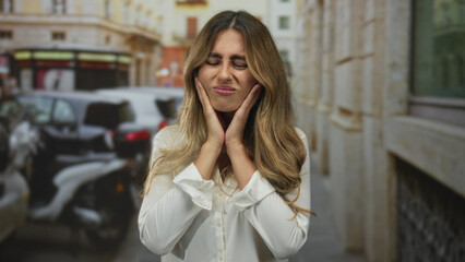 Fototapeta premium Woman with blonde hair in a white blouse holds her cheek with both hands on a narrow city street; toothache discomfort.
