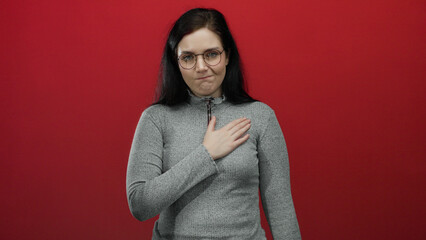 Woman in glasses and gray sweater standing against vibrant red background, looking reflective with...