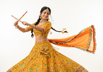A woman in a vibrant yellow and orange traditional outfit garba dances gracefully against a white background. She holds colorful dandiya sticks in each hand.