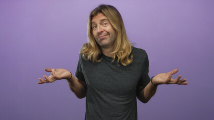 Man shrugs shoulders in studio amid soft purple backdrop with casual grey t shirt and tousled blond...