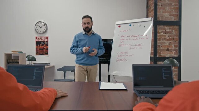 Medium full shot of middle-aged Caucasian male teacher talking to group of multiethnic young prisoners during computer coding lesson, while working on offender rehabilitation program in prison