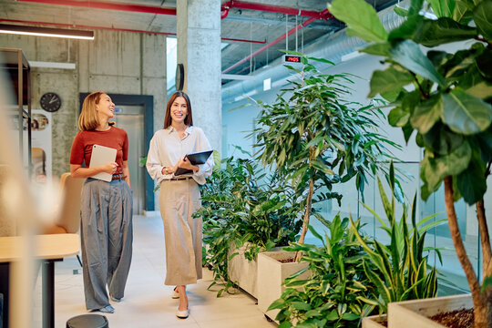 Two happy businesswomen holding tablet are walking through green office space and discussing projects