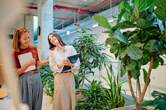 Two businesswomen are walking and talking together holding tablets in a green office space, laughing and enjoying their work