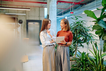 Two businesswomen are standing in a modern office with plants, using a tablet and discussing work