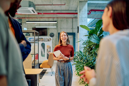 Asian businesswoman holding tablet and smiling while walking towards her colleagues in a modern office, discussing a project