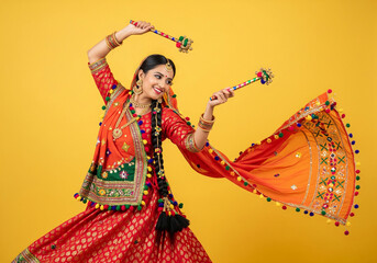 A woman in a vibrant orange and red traditional outfit garba dances gracefully against a bright yellow background. She holds colorful dandiya sticks in each hand.