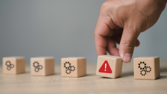 A hand picking a wooden block with an alert symbol among gear blocks, representing issue identification, risk management, and problem detection in process systems.
