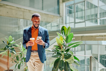 Businessman enjoying a coffee break, smiling while browsing his smartphone in a stylish, modern office filled with bright natural light