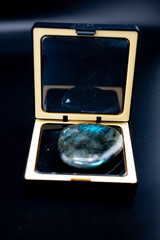 Fototapeta premium Polished labradorite gemstone in a black and gold mirrored compact case on a dark background. Its blue-green iridescence evokes luxury, spirituality, and modern natural decor.