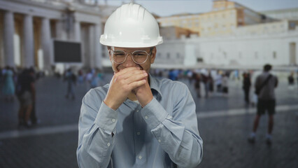 Hispanic man engineer wearing white hardhat holds both hands over mouth in historic building;...