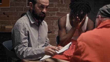 Medium shot of middle-aged Caucasian male prison psychologist talking about forgiveness to convicts at group therapy, while upset young African American inmate is crying, sobbing and praying - Powered by Adobe