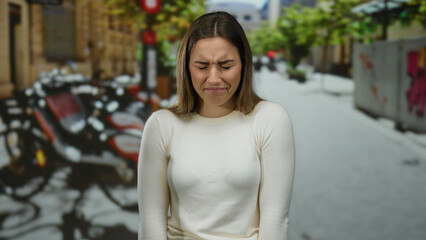 Woman outdoors on a street with blonde hair and neutral expression, white shirt, and blurred city...