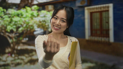 Young chinese woman outdoors holding clipboard while gesturing strength and invitation, surrounded...