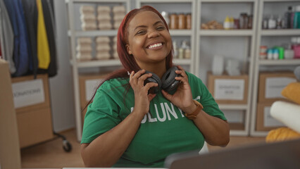 Woman in green volunteer shirt holds headphones to ears and smiles amid stacked donation boxes at indoor center; compassion.