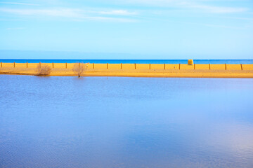 Vivid coastal scene featuring a calm body of water in the foreground and a golden sandy strip leading to deep blue sea. Tranquil coastal landscape with a calm lagoon meeting the horizon