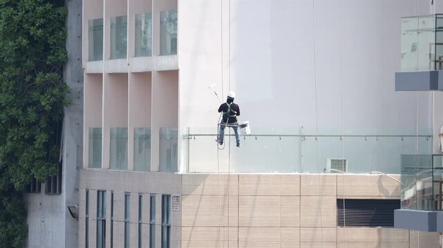 A professional worker with safety equipment paints the tall exterior wall of a modern high-rise building