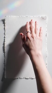 Serene overhead static video, flat lay of pristine handmade white paper with deckled edges and rough texture. A human hand (neutral skin) adjusts it on a soft white background with bokeh, under