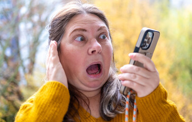 shocked, Surprised Mature woman holds her smartphone and with wide eyes reacts emotionally, looks at screen in shock. Emotional reaction to unexpected news, Viral content, unexpected online event