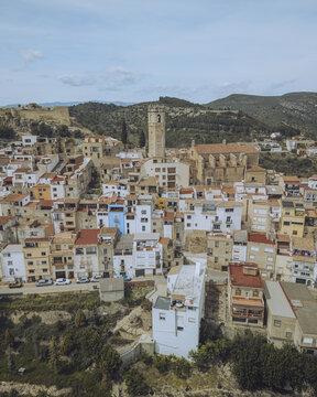 Aerial view of sun-drenched buildings cascade towards a towering church, nestled in a valley beneath ancient castle ruins, Cervera del Maestre, Valencian Community, Spain.
