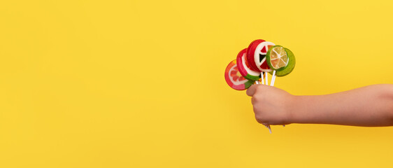 Kid's hand holding a bunch of vibrant fruit-shaped lollipops on sticks against a bright yellow background, evoking fun and sweetness.