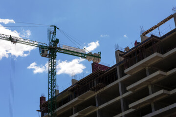 Low-angle view of a concrete building under construction with a green tower crane against a bright blue sky with clouds.