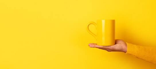 Hand in yellow sweater presenting a yellow ceramic mug on open palm against a solid yellow background.