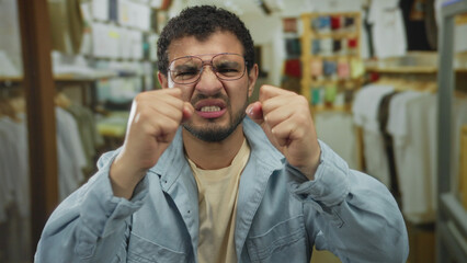 Young hispanic man wearing glasses pressing temples with hands amid clothing racks in retail...