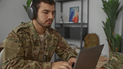 Fototapeta premium Man in camouflage uniform wearing headphones and dog tags typing on laptop in living room; focused discipline.
